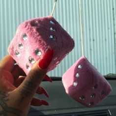 two pink dices are hanging from a car's dash board, and one is being held by a woman with red nails