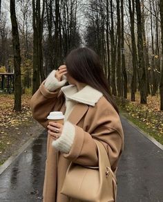 a woman is walking down the street with her coffee in her hand and wearing a camel colored coat