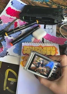 a person holding a cell phone in their hand next to some art supplies and pens