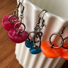 four different colored heart shaped earrings hanging from a metal hook on a white vase next to a wooden table