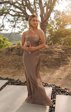 a woman in a long dress holding a wine glass and posing for the camera with a tree behind her