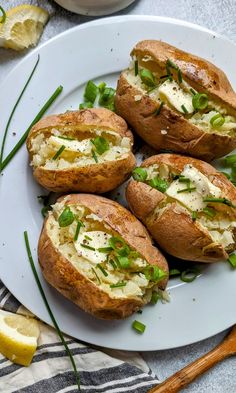 four baked potatoes with cheese and green onions on a white plate next to lemon wedges