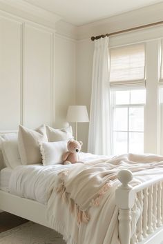 a white bed with pillows and a teddy bear on it in front of a window