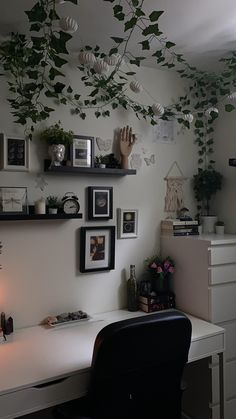 a white desk topped with a black chair next to a wall covered in pictures and plants