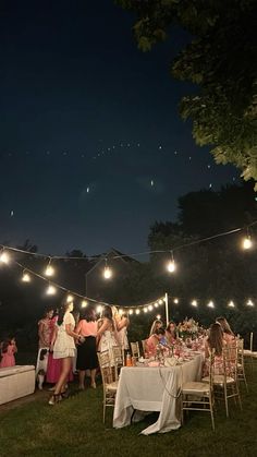 a group of people sitting at tables under string lights in the grass with food and drinks on them