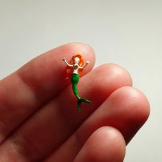 a tiny green and orange toy mermaid sitting on top of a persons finger in front of a white background