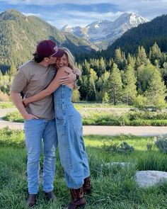 a man and woman hugging each other in the grass with mountains in the back ground