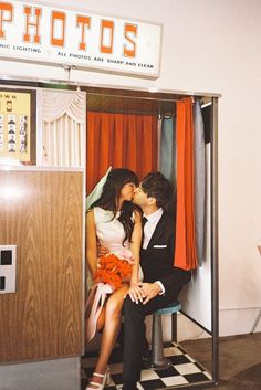 a man and woman sitting on a bench kissing in front of a photo booth with orange drapes