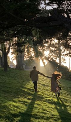 two people holding hands while walking across a grass covered field with trees in the background