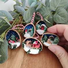 hand holding small christmas ornaments on top of a wooden table next to plants and greenery