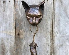 a metal cat head mounted to the side of a wooden wall with a rat hanging from it's tail