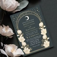 the wedding card is next to some white flowers and greenery on a black surface