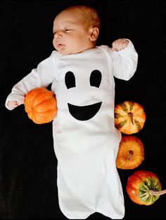 a baby dressed in a ghost costume laying next to pumpkins and apples on a black background