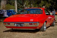 an old red car is parked on the grass near other cars and people in the background