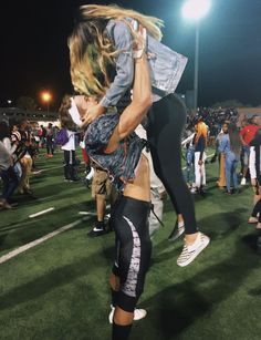 a woman holding onto another woman on top of a football field at night with other people in the background