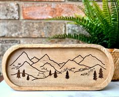 a wooden tray with mountains and trees on it next to a potted fern plant