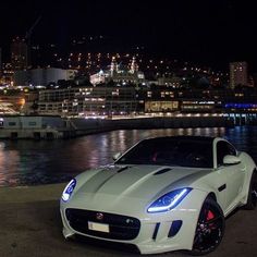 a white sports car parked in front of the water at night with city lights behind it