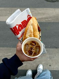 a person holding a cup of soup and a sandwich in their hand on the street