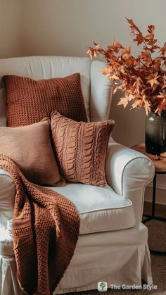 a white chair with some pillows on it and a vase filled with flowers in the corner