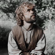 a man with curly hair and beard wearing a brown vest sitting in front of trees