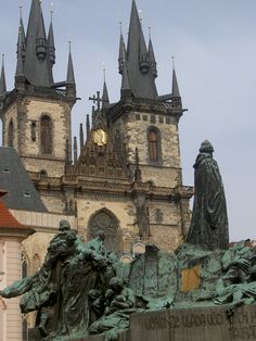 a statue in front of a large building with towers on it's sides and a clock at the top