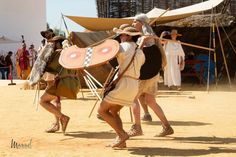 some people are dressed up and dancing in the sand while others stand around with shields on their heads