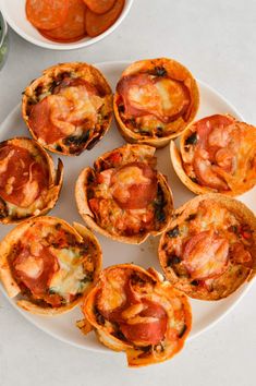 several mini pizzas on a white plate next to bowls of sauce and pepperoni