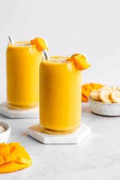 two glasses filled with orange smoothie on top of a white counter next to bananas