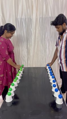 two people standing next to each other near a black table with green and blue cups on it