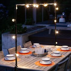 an outdoor dining table set with plates and cups on it, lit by string lights