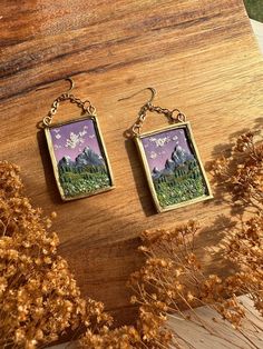 two small square earrings with mountains in the background on a wooden table next to dried flowers