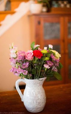 a white vase filled with lots of colorful flowers on top of a wooden table next to a stair case