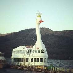 a large white duck floating on top of a lake next to a boat dock and mountains in the background