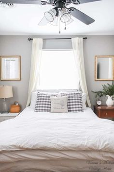 a white bed sitting under a ceiling fan in a bedroom next to a window with two framed pictures on the wall