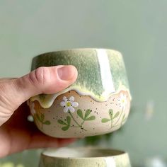 a hand holding a green and white cup with flowers painted on the outside, in front of a wall