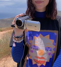 a woman holding a camera up to her face while standing on top of a hill