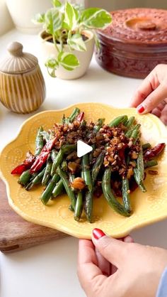 two hands are holding a yellow plate with green beans and other food items on it