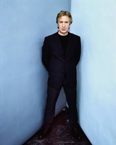 a man in a black suit leaning against a wall with his hands on his hips