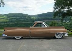 an old brown car parked in a parking lot next to some trees and hills behind it