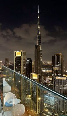 a balcony overlooking the city at night with chairs and tables set up on it's glass railing