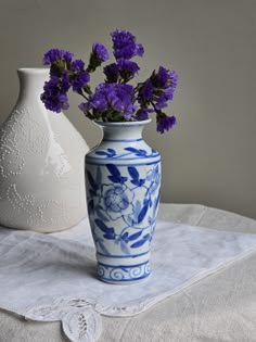 two vases with purple flowers in them on a white table cloth next to each other