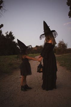 two children dressed up as witches holding hands on a dirt road at dusk with trees in the background