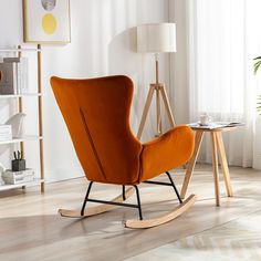 an orange rocking chair sits in front of a white wall and wooden table with a lamp on it