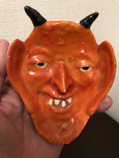 a hand holding an orange ceramic mask with horns on it's head and eyes