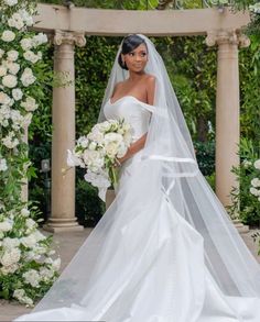 a woman in a wedding dress holding a bouquet and standing under an arch with white flowers