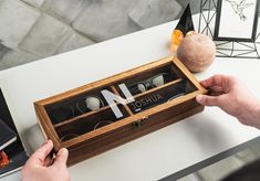 two hands holding an open wooden box with glasses in it on a table next to other items