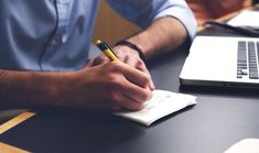 a man is writing on a piece of paper while sitting at a desk with a laptop