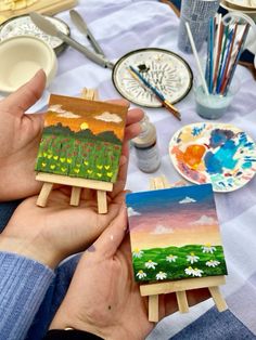 a person holding two small paintings on easels in front of some paintbrushes