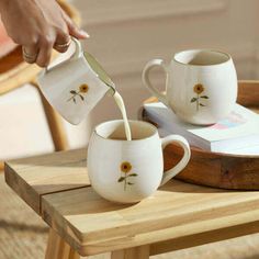 a person pouring milk into two mugs on a table with a book and chair in the background