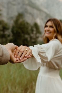 a man and woman holding hands while standing in tall grass with mountains in the background